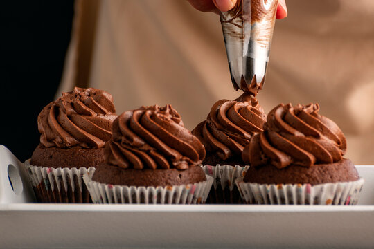 Pastry chef decorates cupcakes with chocolate cream from pastry bag. Homemade cocoa muffins close up.