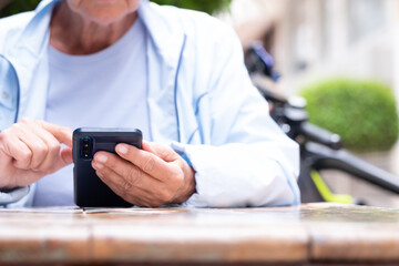 Caucasian woman holding in hands and using smart phone while sitting at an outdoor table. Elderly woman sitting near her electric bike enjoying tech and social
