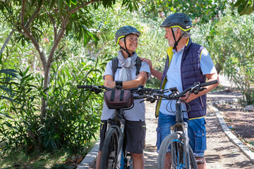 Active senior couple with electric bicycles standing outdoors in the park looking  each other while chatting. Two smiling seniors wearing cycling helmet in the nature enjoying a healthy lifestyle