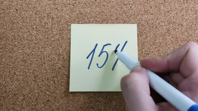 Handwritten inscription "15%" in blue marker on a yellow paper sticker. Sticker on a cork board close-up. Blue felt-tip pen in a woman's hand