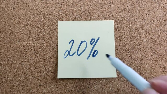 Handwritten inscription "20%" in blue felt-tip pen on a yellow paper sticker. A blue marker in a woman's hand. Sticker on a cork board close-up