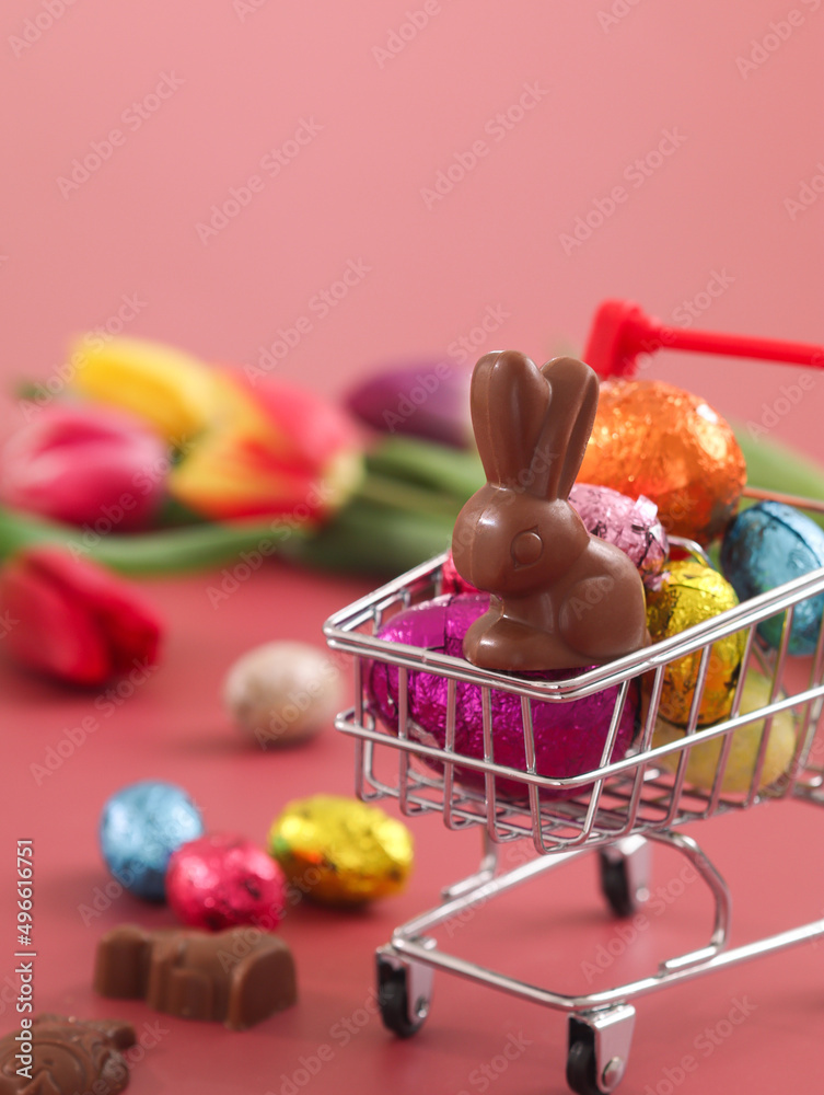 Wall mural Close up of Easter rabbit chocolate in a mini shopping cart with colorful Easter egg, and festive decoration atmosphere. Colorful tulips flower with pink background