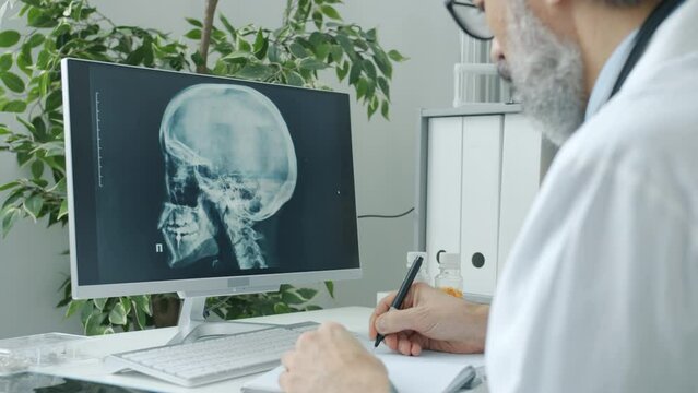 Doctor radiologist analyzing human skull images looking at computer screen and witing indoors in hospital. Modern technology and radiology concept.
