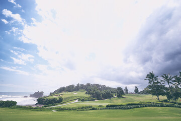 Beautiful balinese landscape. Golf field on ocean shore. Ancient hinduism temple Tanah lot on the rock against cloudy sky. Bali Island, Indonesia.