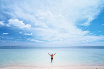 Fototapeta na wymiar Vacation and freedom. Happy young man rising hands up standing on tropical beach enjoying beautiful view.