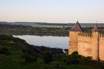 On the right side of the tower of a medieval stone fortress with battlements. On the left is a river with banks in the grass