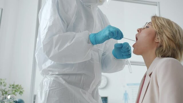 Doctor in protective suit testing female patient for corona virus infection in hospital laboratory taking PCR throat swab in test tube