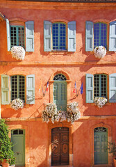 View on traditional colorful ochre natural stone wall house facade, flower arrangements, old window wood shutters - Roussillon town hall, France