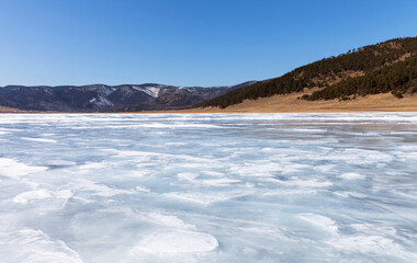 Fototapeta na wymiar Baikal Lake in early spring on sunny day. Ice surface was covered with thawed patches and snow on ice became loose and wet. Coastal hill cleared of snow. Beautiful spring landscape. Natural background