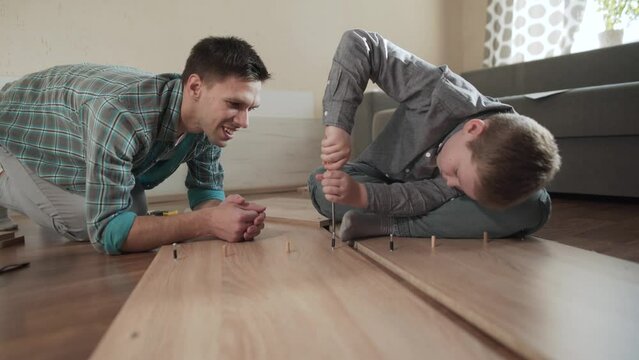 Caucasian son sits on chipboard piece and screws a screw with screwdriver. Young dad carefully watches work of his son and helps to screw the mount evenly. Family assembles furniture together.