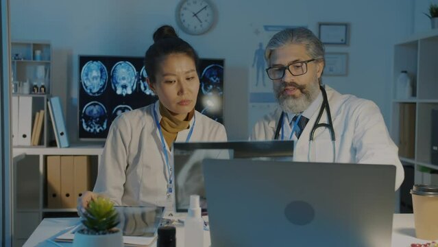 Asian woman and Caucasian man doctors analyzing lungs x-ray image and talking working in hospital at night. Medicine and corona virus pandemic concept.