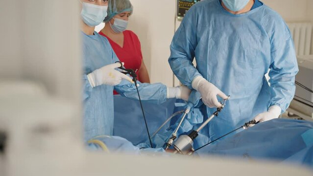 Portrait of female medical worker in protective mask and sterile gloves holding laparoscopic grasper with trocar while standing near table with surgical tools. Surgeon and his assistant on blurred.
