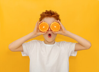 A red-haired boy holds in front of his eyes the halves of a juicy orange on a yellow background