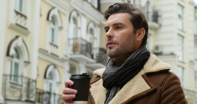 Handsome man in suit is holding a cup of coffee and smiling while walking outdoors. cheery male drinking takeaway coffee while standing over building. stylish man in a jacket wait for a girl on a date
