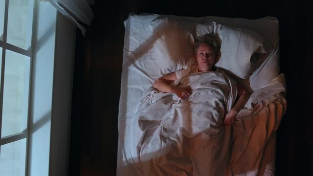 Annoyed woman can't sleep because of loud music while lying in bed in bedroom at night, top view. Sleepy person experiences anger, irritable and  hostile mood.