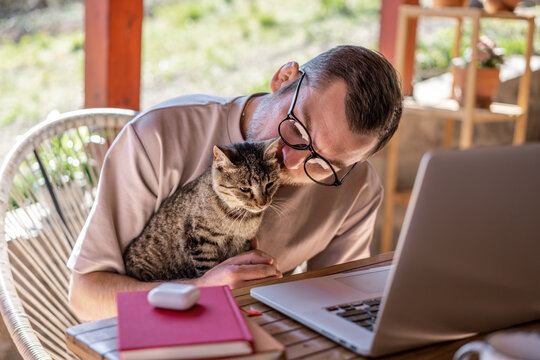 Young attractive man in glasses freelancer with a gray cat in his hands working at home using a laptop on the terrace of a country house