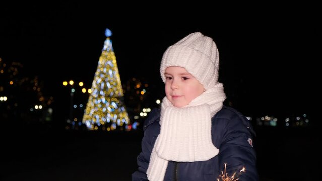 A beautiful Christmas child plays with a sparkler in front of a Christmas tree in the evening outdoors.