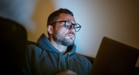 Young attractive serious man in glasses sitting at home at night on an armchair looking into a...