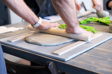 A close-up of a man potter rolls a brown clay rolling pin on a special fabric on a wooden table to make a plate