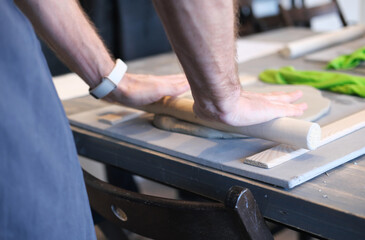 A close-up of a man potter rolls a brown clay rolling pin on a special fabric on a wooden table to make a plate