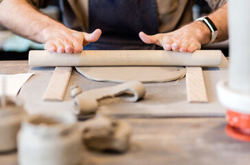 A close-up of a man potter rolls a brown clay rolling pin on a special fabric on a wooden table to make a plate