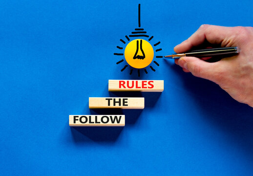 Follow The Rules Symbol. Concept Words Follow The Rules On Wooden Blocks. Businessman Hand. Yellow Light Bulb Icon. Beautiful Blue Background. Business And Follow The Rules Concept. Copy Space.