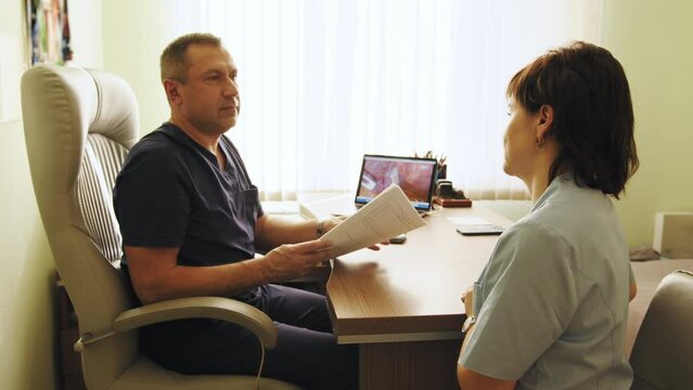 Two doctors holding and talking about a file. Doctors discussing and working together in a medical office. Doctors talking in an hospital. Colleagues in medical uniform sitting by desk in office.