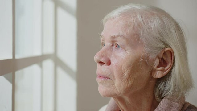 Elderly woman of 80 years old with sad eyes looks out window portrait. Concept of loneliness in old age.