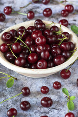 Freshly gathered cherries with leaves and stalks in a plate and on grey surface background inside, fresh red cherries in dish and table, harvest of red cherries in rustic style