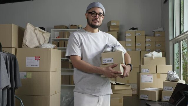 Confident young Asian man retail seller, entrepreneur, online store dropshipping small business owner looking at camera standing in delivery shipping warehouse with parcel boxes.