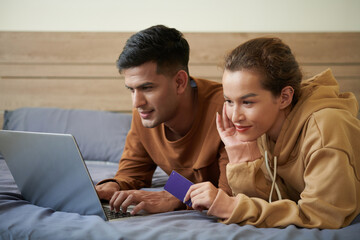 Cheerful young couple relaxing on bed and shopping in favorite online store enjoying seasonal sale