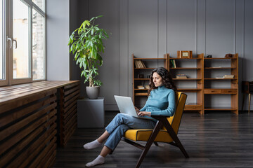 Young woman freelancer work on computer at home sit in armchair typing email to partner colleague....