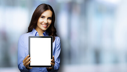 Portrait of smiling brunette businesswoman showing mock up blank tablet pc, touchpad, blurred modern office interior background. Confident business woman indoors.