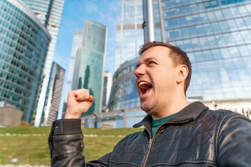 Millennial excited man celebrating his success standing outdoors in urban area. Cheerful handsome male with arms raised in air, celebrating victory standing outside office building. Winner and achieve