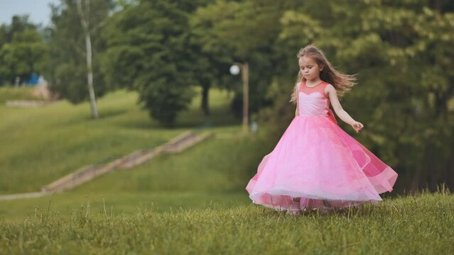 A girl in a pink princess dress is spinning in the park.