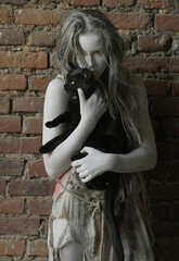 A girl is dressed in rags as a Halloween horror 
figure. She stands in front of an old brick wall. Her 
clothes are torn and dirty and she holds a black cat 
in her arms.
