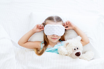 a little girl child with a sleep mask woke up in the morning at home on a bed on a white cotton bed with a teddy bear toy and a cute smile