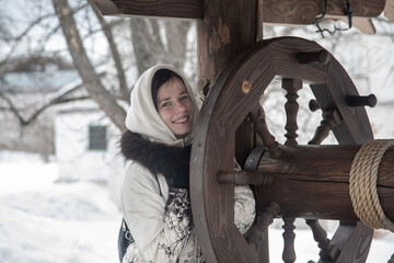 Fototapeta na wymiar A beautiful girl is sitting by the well. Old wooden well. The girl is sitting and smiling. Winter cloudy snowy day.