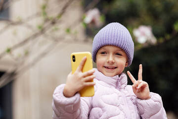 Cute little caucasian girl with blonde hair in trendy casual clothes using mobile phone outdoor at spring. Video chat and social distancing during coronavirus covid-19 pandemic lockdown
