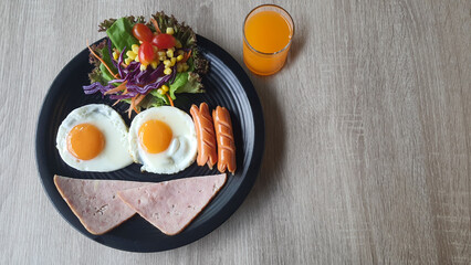 Top view of fried egg and sausage and ham smiley face and colorful salad like hair on black plate and orange juice at wood table, breakfast serve for kid, funny breakfast.
