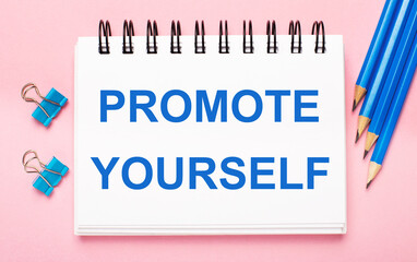 On a light pink background, light blue pencils, paper clips and a white notebook with the text PROMOTE YOURSELF