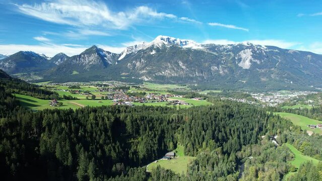 High view of a beautiful Austrian village surrounded by green landscape with snow covered mountain tops and blue sky.