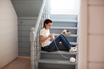 Calm woman knit white thread on knitting needles, sitting on steps in cozy wooden interior house....