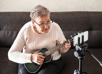 Elderly woman 70+ sits on the couch and learns to play the ukulele at home using video tutorial on her smartphone. Adaptation of pensioners in the modern world. New knowledge and skills. Hobby