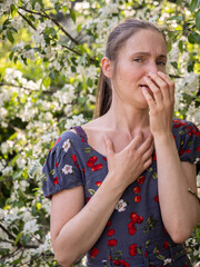 Young woman sneezes in the park against the background of a flowering tree. Allergy to pollen concept.
