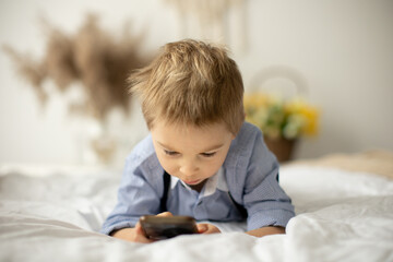 Blond preschool child, cute boy, playing on mobile phone in bed