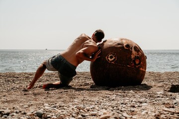 Man doing exercise outdoors near to old rusty floating marine mine on the beach with rocky shore...