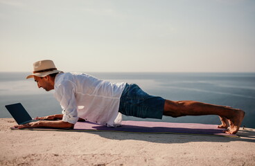 A digital nomad, a man in a hat, a businessman with a laptop does yoga on the rocks by the sea at sunset time, does a business operation online from a distance. Remote work on vacation.