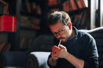 Smoking Male student wearing glasses in library with bookshelves on background.