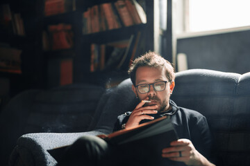 Adult student reading a book in the college library while smoking. Young male wearing glasses with...